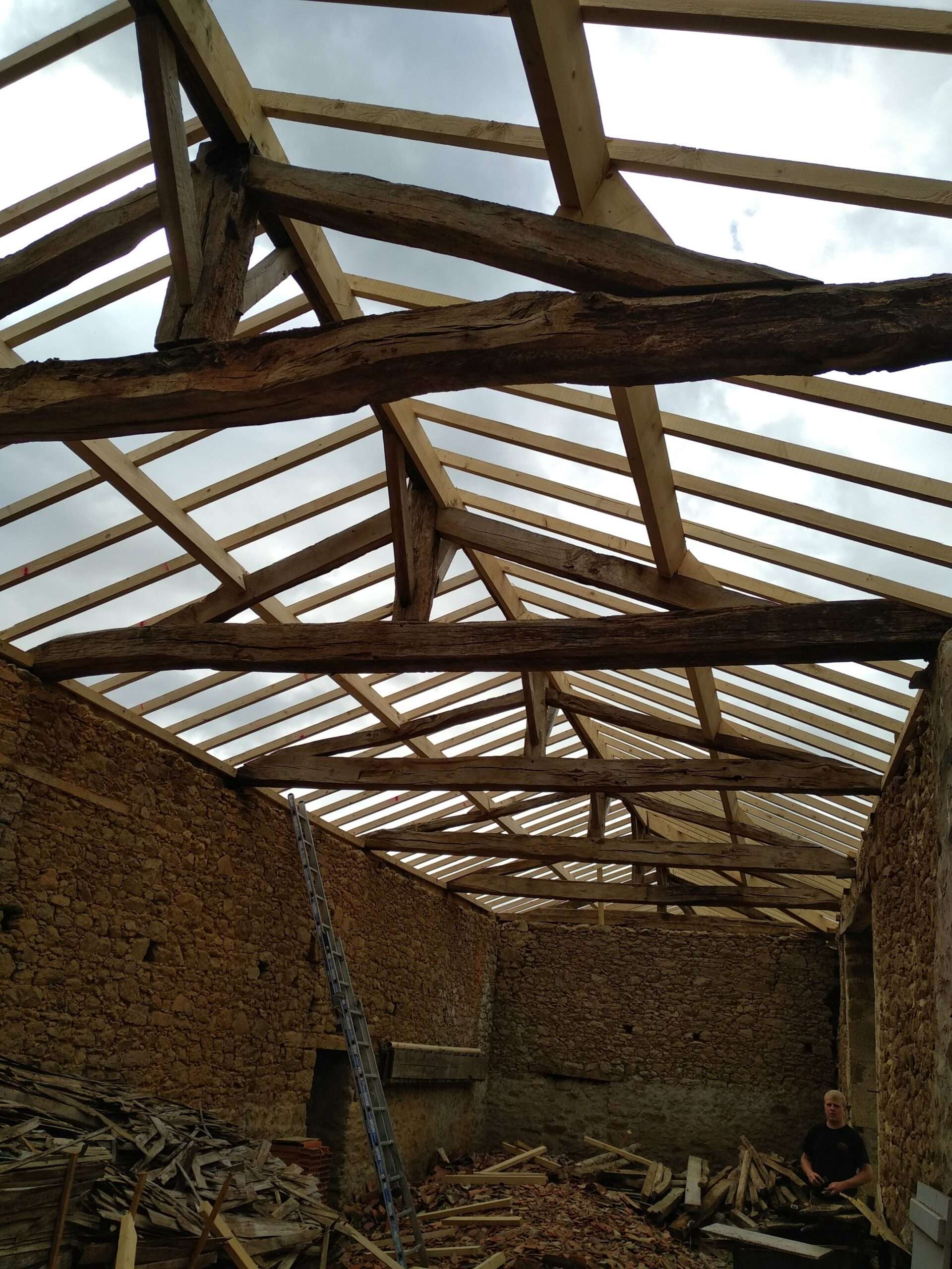 Une charpente en bois partiellement restaurée à l'intérieur d'un bâtiment en pierre ; une échelle est appuyée contre le mur, et une personne est assise au milieu de débris de chantier éparpillés.