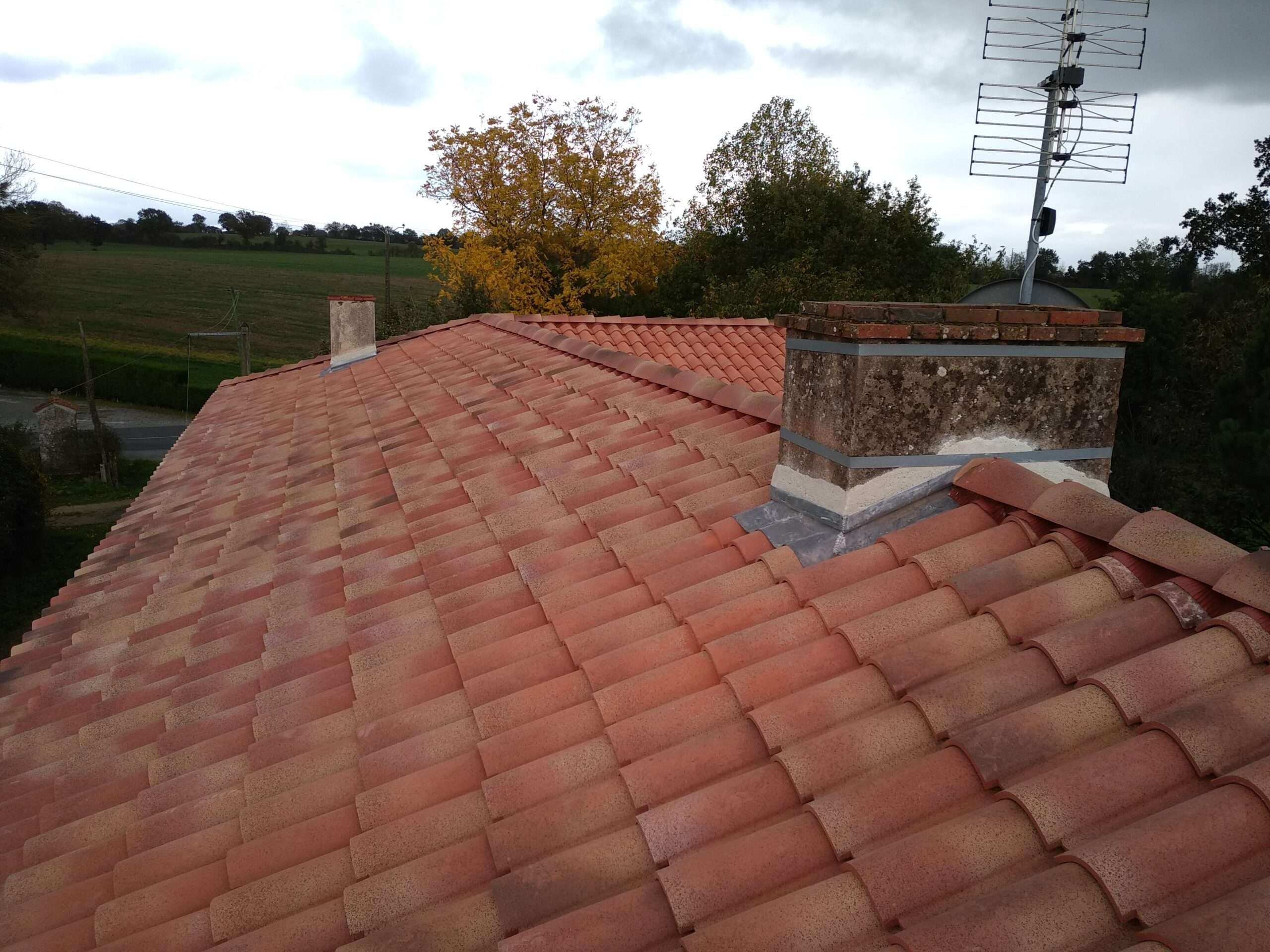 Toit en tuiles d'argile rouge posé de main de maître par un couvreur professionnel, doté de deux cheminées et d'une antenne de télévision, avec des arbres et un champ visibles à l'arrière-plan sous un ciel nuageux.