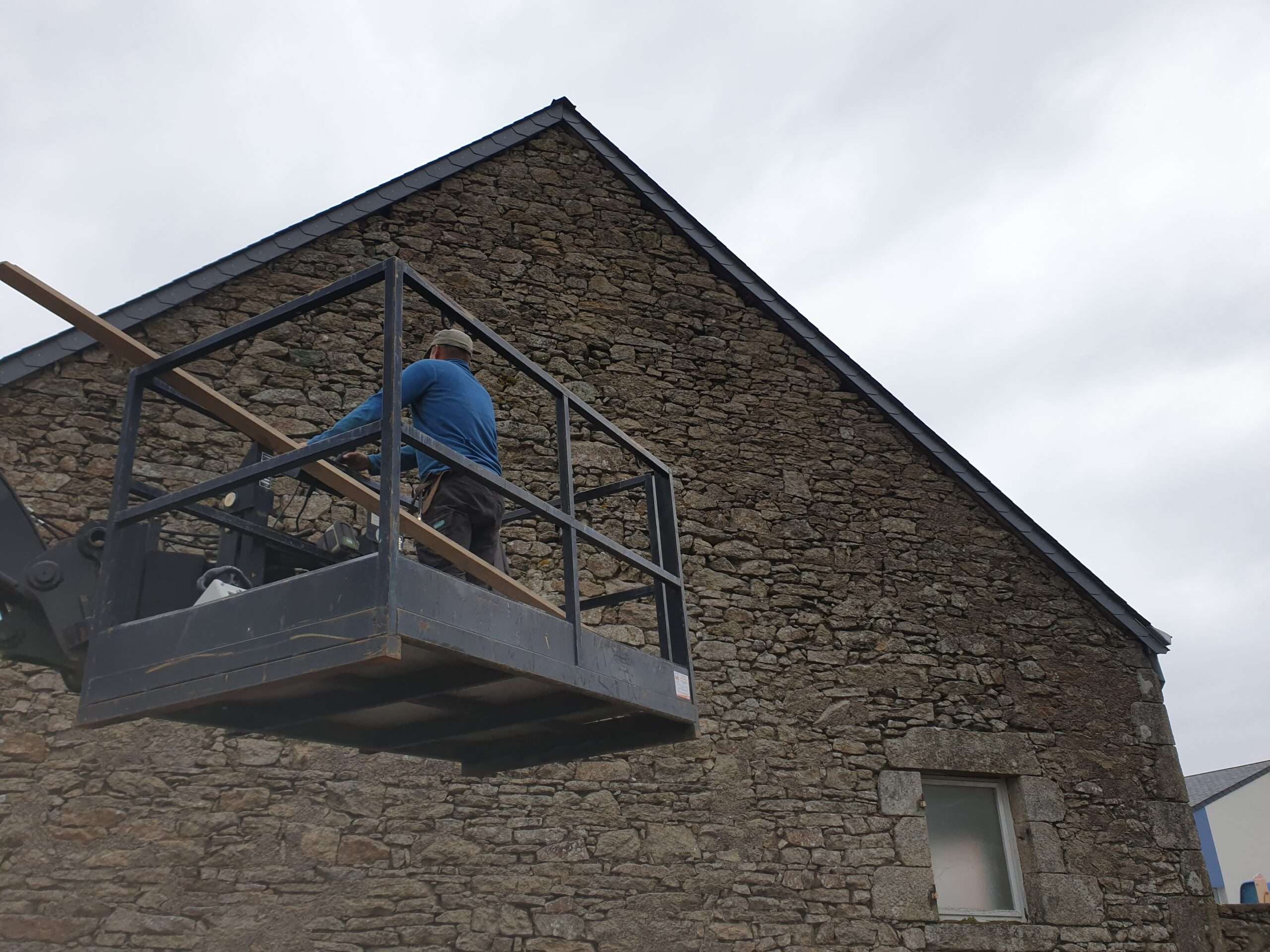 Une personne se tient sur une plate-forme de travail surélevée près d'un bâtiment en pierre, tenant une longue poutre en bois, par une journée nuageuse.