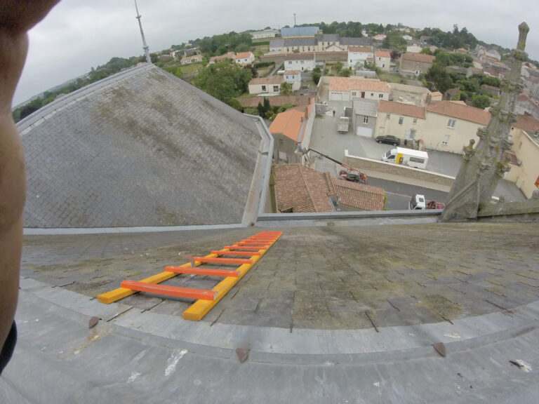 La vue depuis un toit montre une échelle orange, des maisons de village, des véhicules garés et de la verdure en arrière-plan, sous un ciel nuageux.