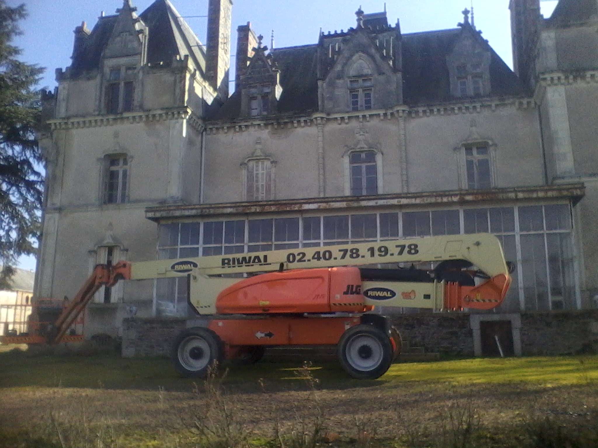 Une nacelle élévatrice mobile est garée devant un vieux bâtiment en pierre richement décoré, doté de toits en pointe et de grandes fenêtres.