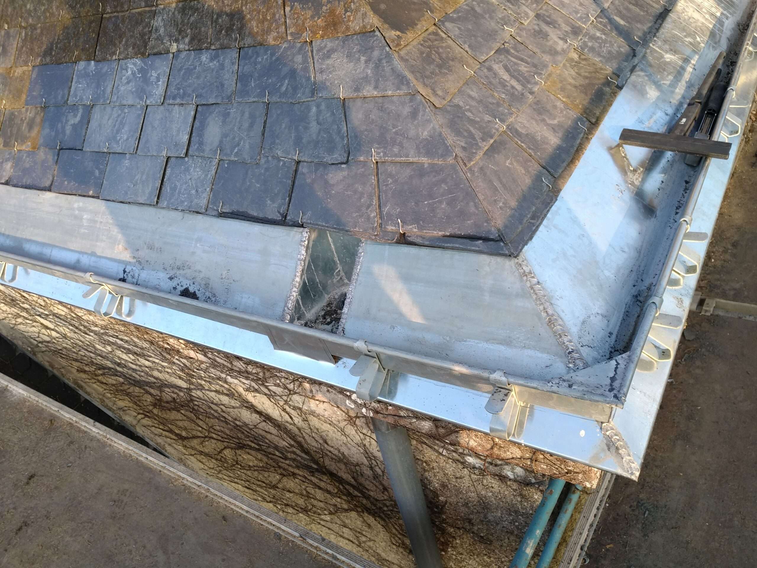 Gros plan sur l'angle d'un bâtiment avec des tuiles en ardoise, des solins métalliques et un système de gouttières, montrant les traces d'altération et les supports de fixation.