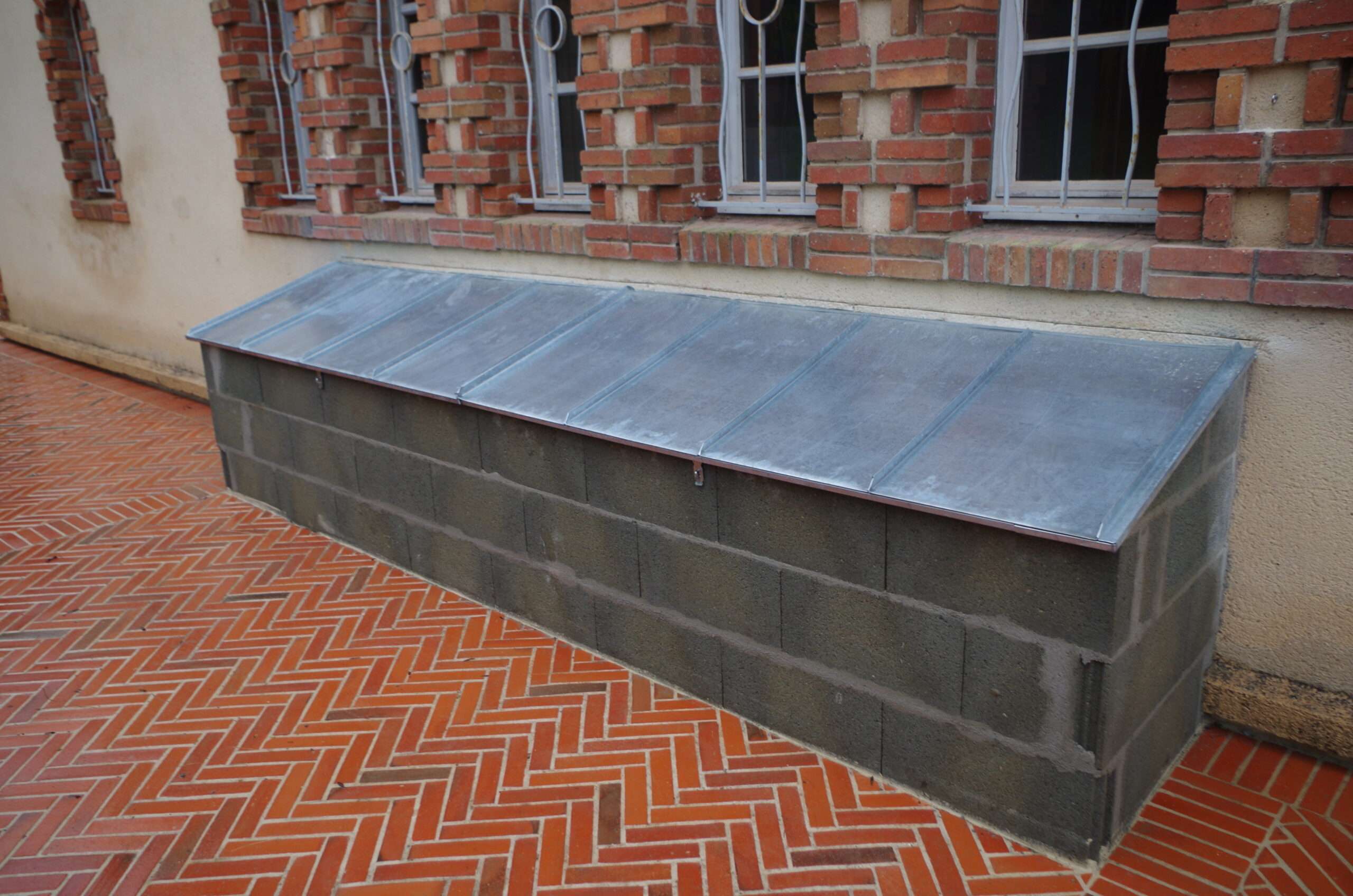 Structure rectangulaire en béton recouverte d'une toiture métallique en pente, adossée à un mur de briques surmonté de plusieurs fenêtres, sur une terrasse carrelée.