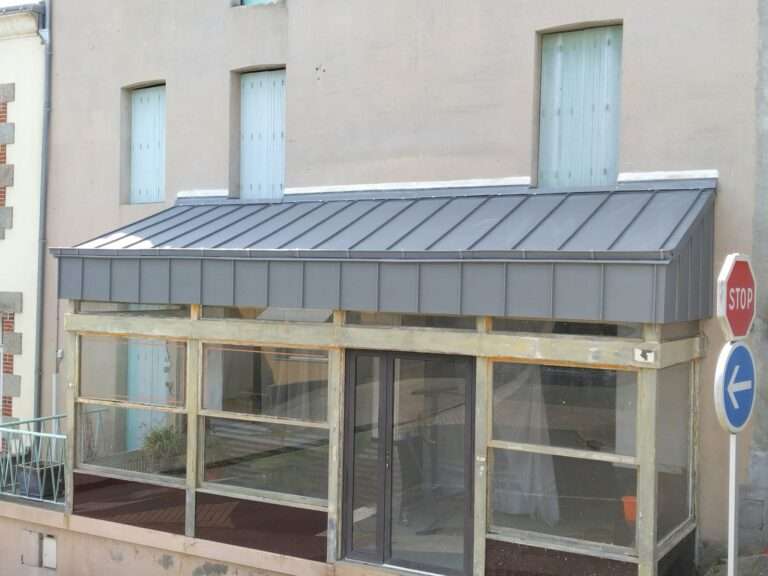 Vitrine avec une toiture métallique recouverte de zinc, adossée à un bâtiment beige à Pouzauges ; les fenêtres situées au-dessus sont masquées, et on aperçoit des panneaux « Stop » et « Tour à droite ».