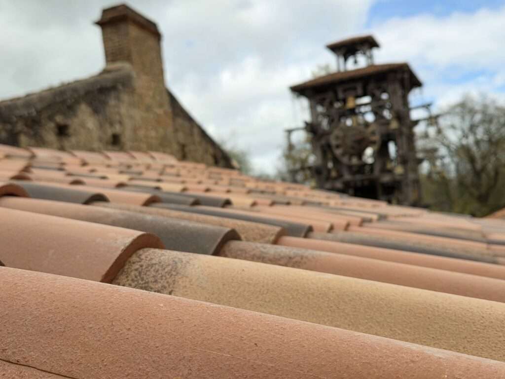 Gros plan sur des tuiles en terre cuite, récemment restaurées dans le cadre d'une réfection de toiture en Vendée, avec en arrière-plan, flou, un bâtiment en pierre rustique et un clocher en bois, sous un ciel partiellement nuageux.