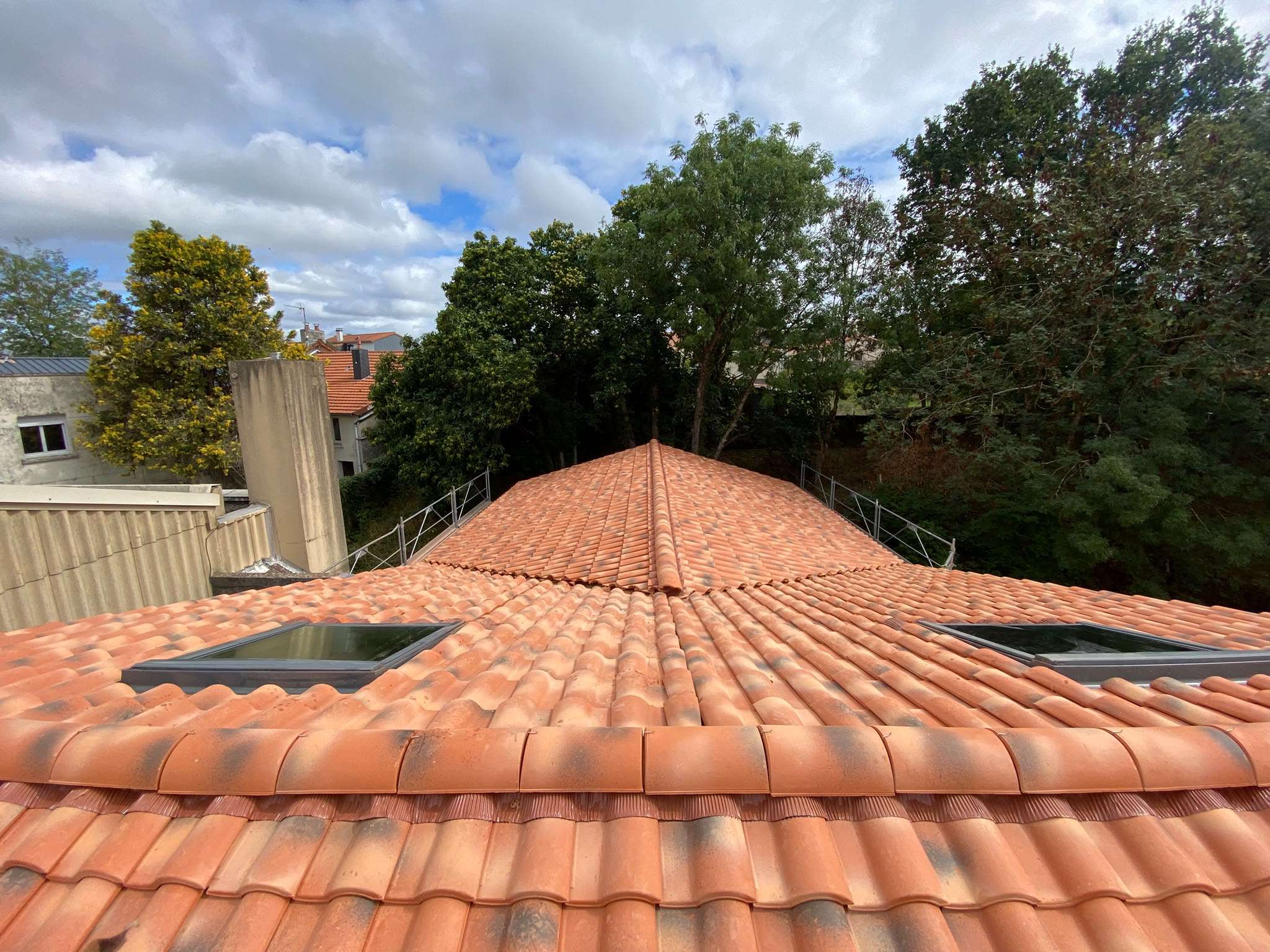 Vue d'un toit recouvert de tuiles rouges, doté de deux lucarnes, entouré d'échafaudages et d'arbres, sous un ciel partiellement nuageux — une scène qui laisse supposer que des travaux de réparation sont en cours, peut-être en raison d'une fuite de toiture.
