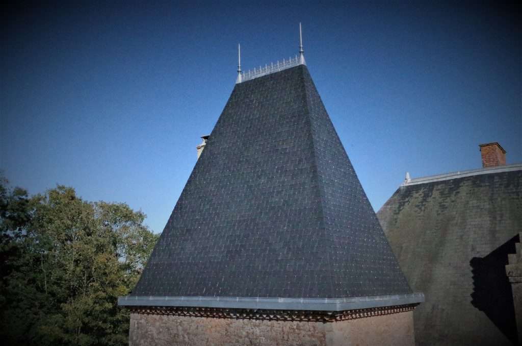 Toit en ardoise à forte pente, de forme pyramidale, surmonté de quatre fleurons métalliques, réalisé avec savoir-faire par l'entreprise de couverture Vendée, adossé à un bâtiment en briques, sur fond de ciel bleu limpide.