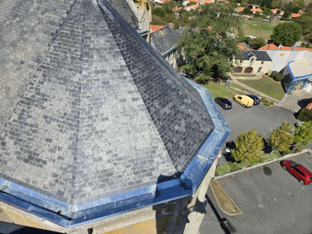 Vue aérienne du toit en ardoise d'une église, où l'on distingue des travaux de réfection et des chenaux en zinc, à côté d'un parking où sont garées deux voitures ; des arbres et des bâtiments voisins sont visibles à l'arrière-plan.