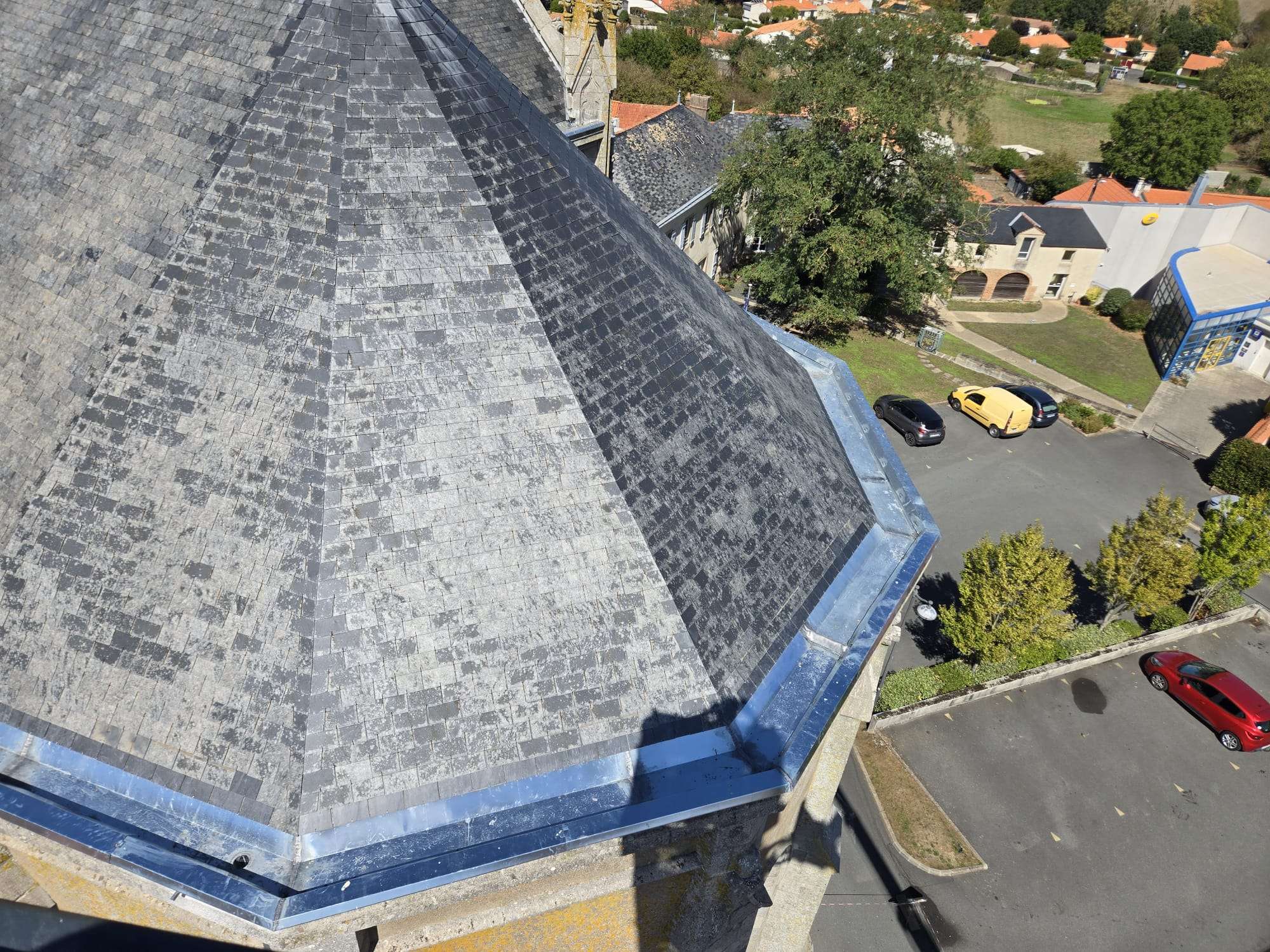Vue aérienne du toit en ardoise d'une église, où l'on distingue des travaux de réfection et des chenaux en zinc, à côté d'un parking où sont garées deux voitures ; des arbres et des bâtiments voisins sont visibles à l'arrière-plan.