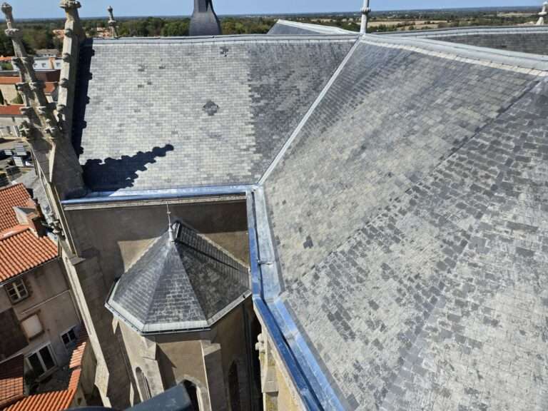 Gros plan sur le toit d'une grande église recouvert d'ardoises grises, avec ses faîtes entrecroisés et sa petite tourelle, sur lequel la rénovation des gouttières en zinc vient d'être achevée ; on aperçoit les bâtiments voisins et le paysage au loin.